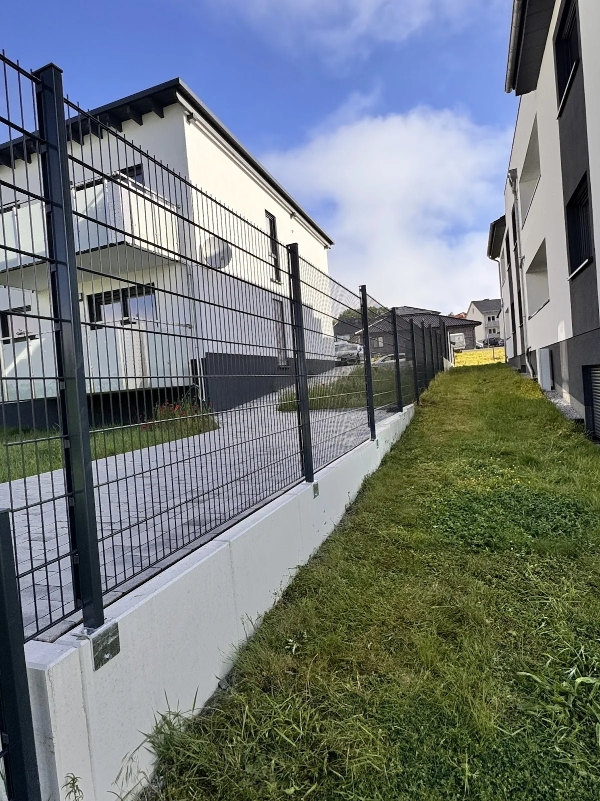 Doppelstabmattenzaun auf weißem Betonsockel neben einem Mehrfamilienhaus