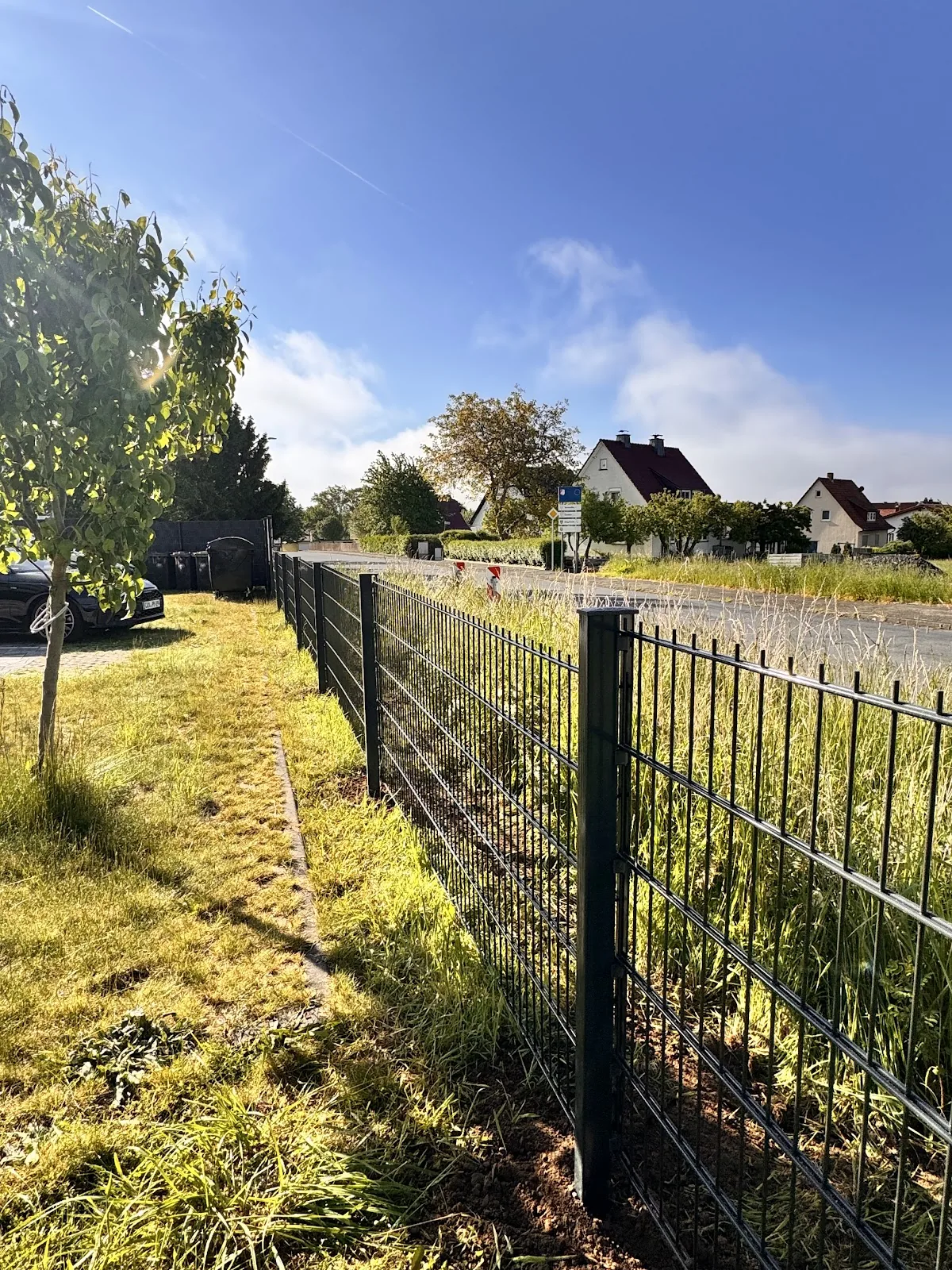 Zaun entlang einer Wiese am Feldweg, dahinter Wohnbebauung